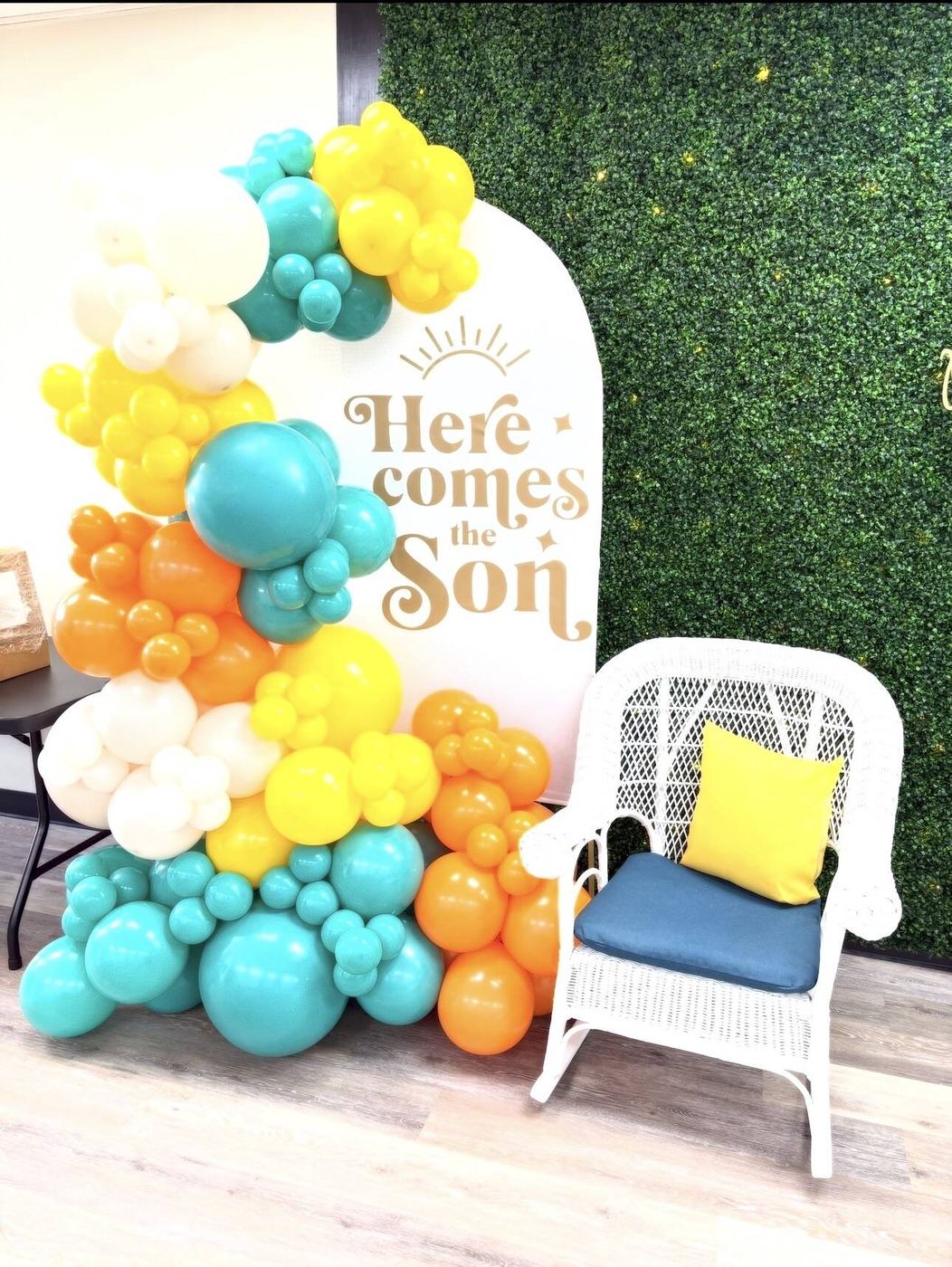 Teal, orange and yellow balloon garland on a Here Comes the Son arch backdrop beside a white wicker chair